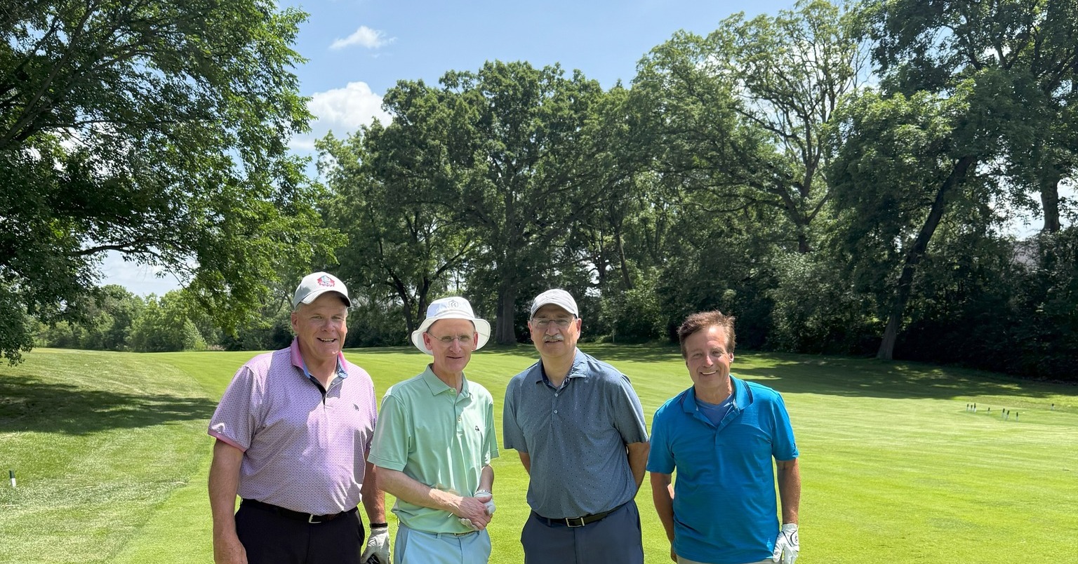 Four men stand on a golf course on a bright sunny day